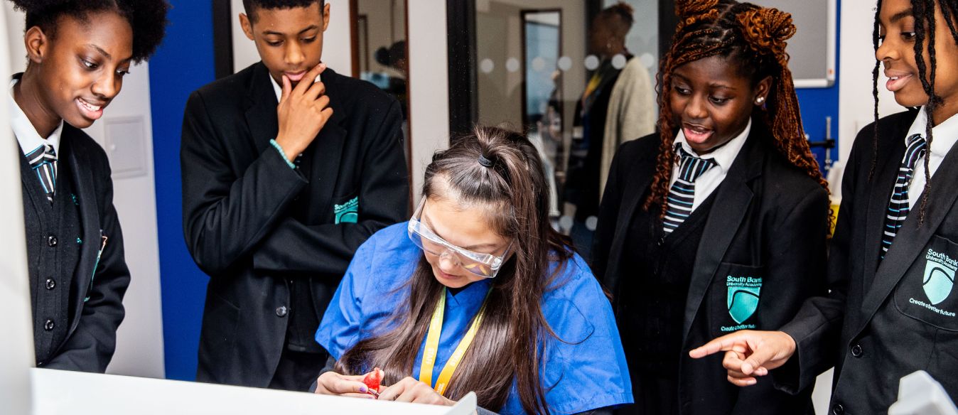 Students in uniforms watch intently as a classmate in safety goggles and a blue lab coat performs an experiment. The scene is focused and educational.