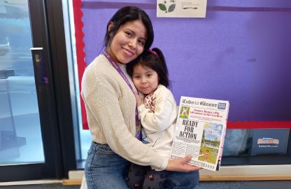 A Lambeth College adult female student gently holds her child in front of a vibrant purple curtain at the Lambeth College Nursery, showcasing a warm and intimate moment between them. The lady is holding a personalised newsletter titled 
